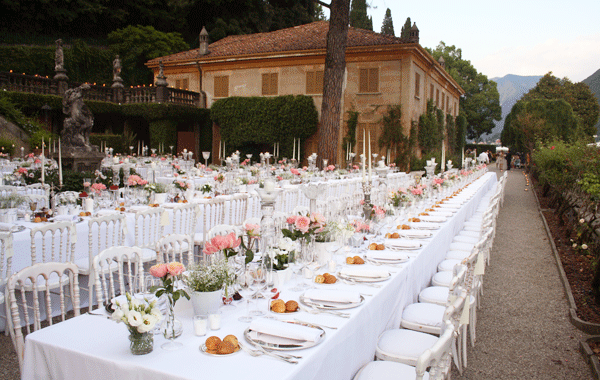 Long outdoor banquet table decorated with pink and white flowers, set with white plates, silverware, napkins, and bread rolls, in a garden setting with lush greenery and a stone building in the background.
