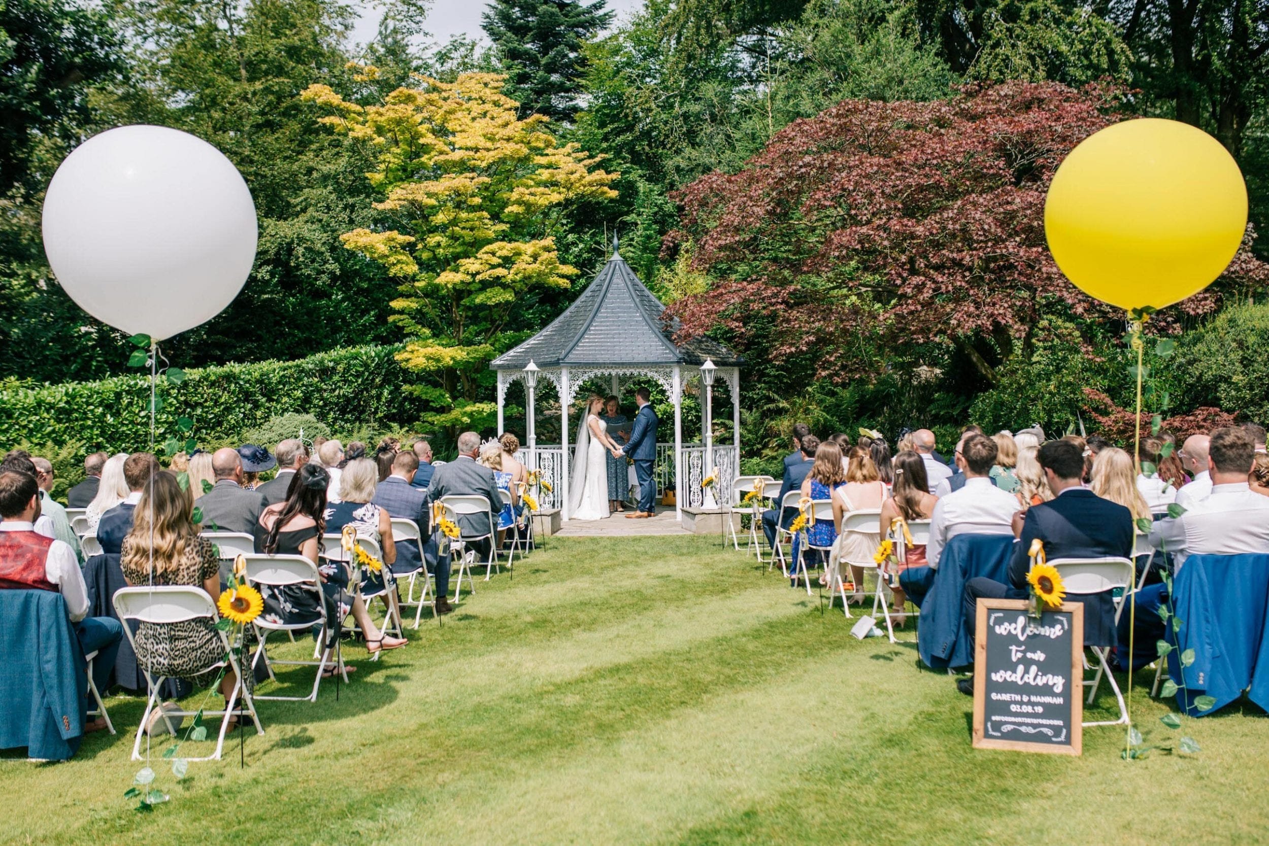 An outdoor wedding ceremony in a garden setting with guests seated on white foldable chairs. A bride and groom stand at a gazebo in the background. Large white and yellow balloons are tied to the chairs along the aisle. There are sunflowers and a wed