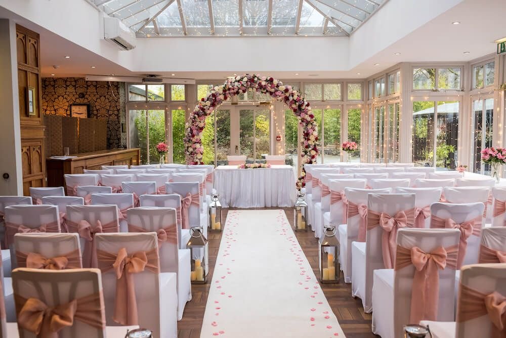 Indoor wedding ceremony setup with decorated chairs and floral arch.