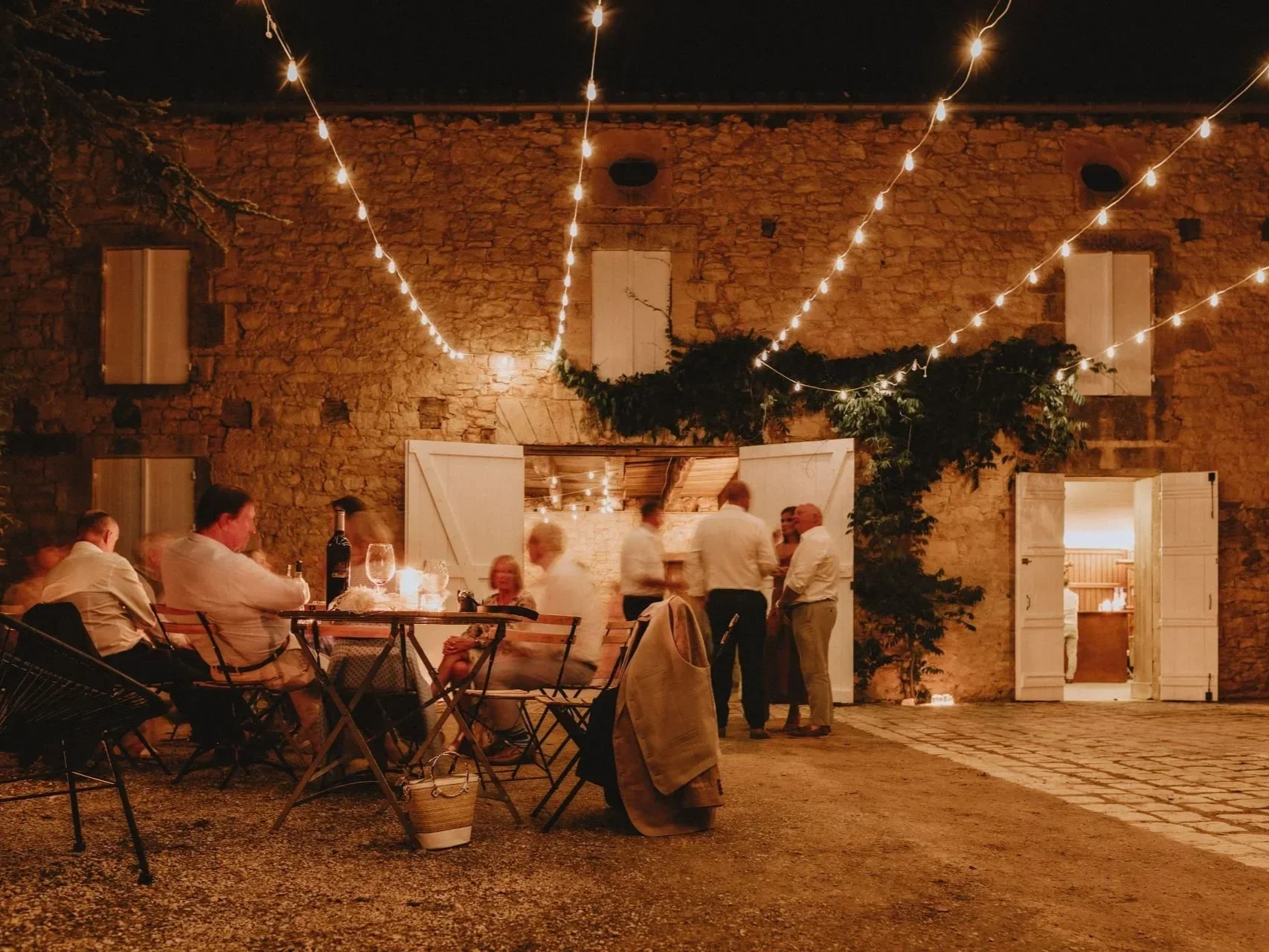 Nighttime outdoor gathering with people socializing under string lights near a rustic brick building with open white-wooden doors, and tables with wine glasses and bottles.
