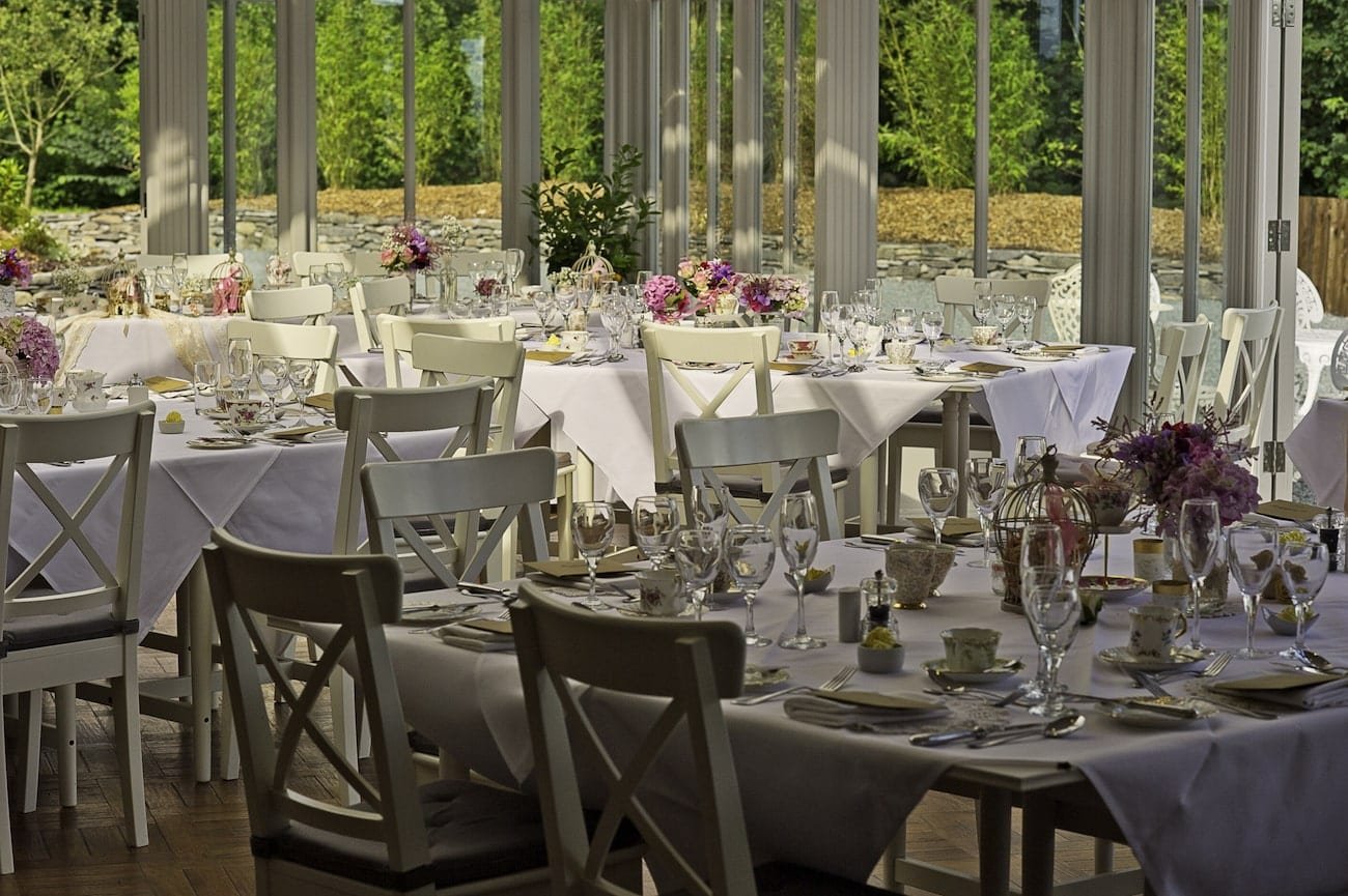 Elegant dining setup with white tables and chairs, decorated with floral centerpieces, glassware, and tableware in a well-lit restaurant or event space.