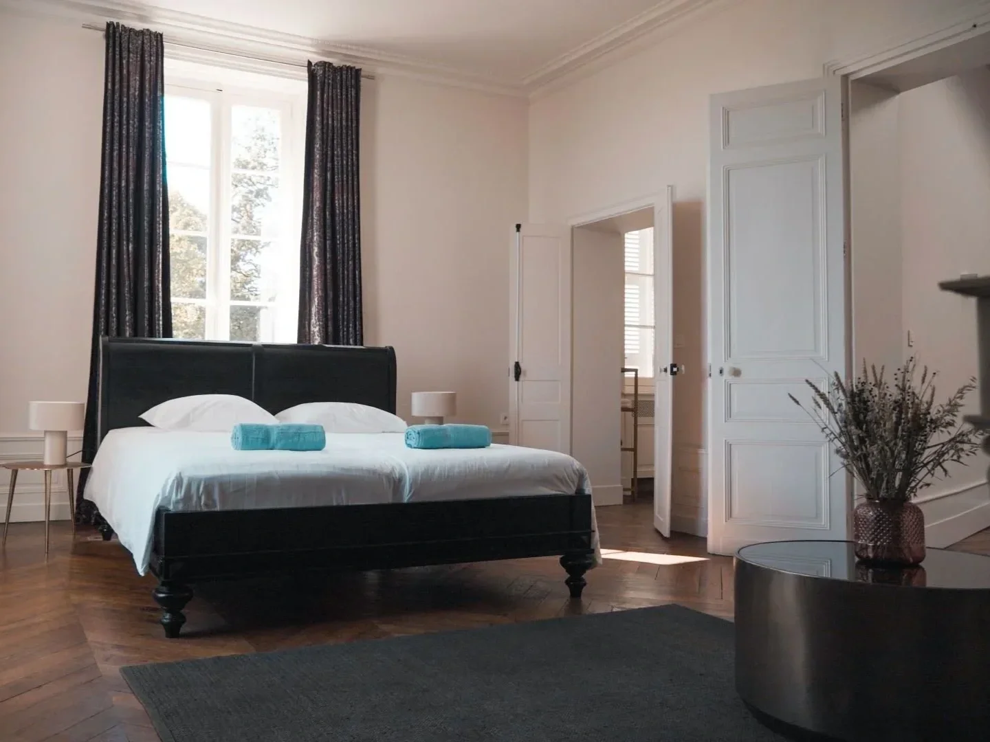 A bedroom with a large window, a black bed frame with white bedding and two rolled blue towels, two small side tables with lamps, a dark rug, a round black coffee table with a vase of dried flowers, and open white doors leading to other rooms.