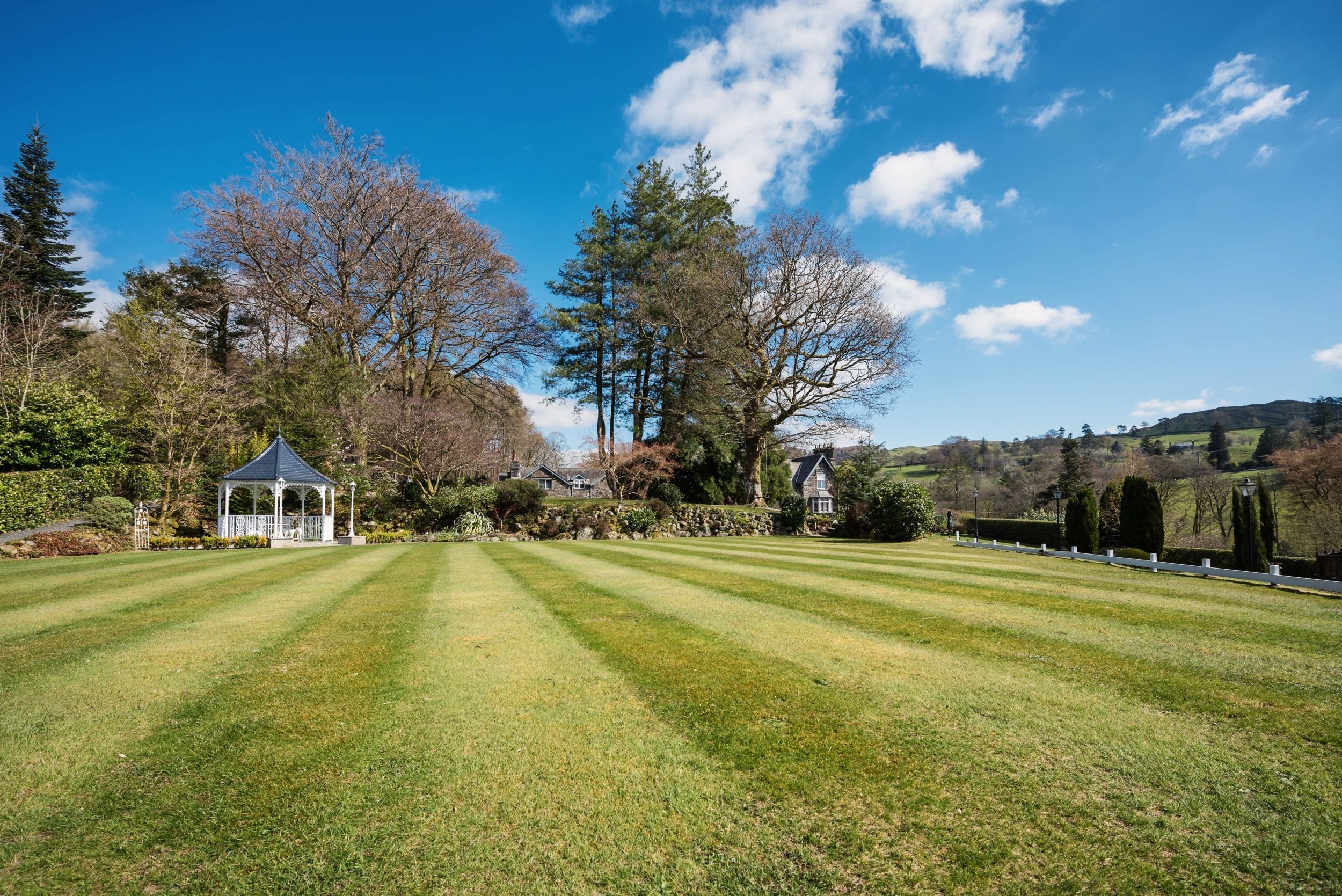 A scenic landscape with a well-maintained lawn in front of a gazebo, surrounded by trees and shrubs, with a house in the background under a blue sky with clouds.