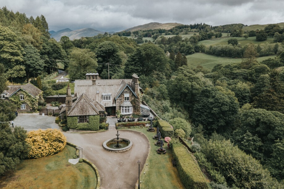Aerial view of a large estate house with a fountain in the driveway, surrounded by lush greenery and hilly landscape under a cloudy sky.