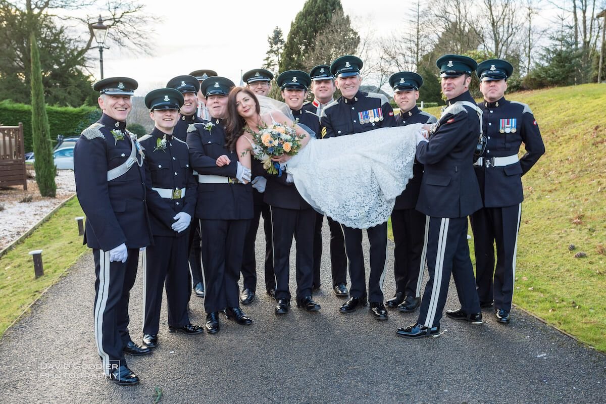 A group of uniformed military personnel holding a bride in a white gown and veil, with a bouquet of flowers, in an outdoor setting.