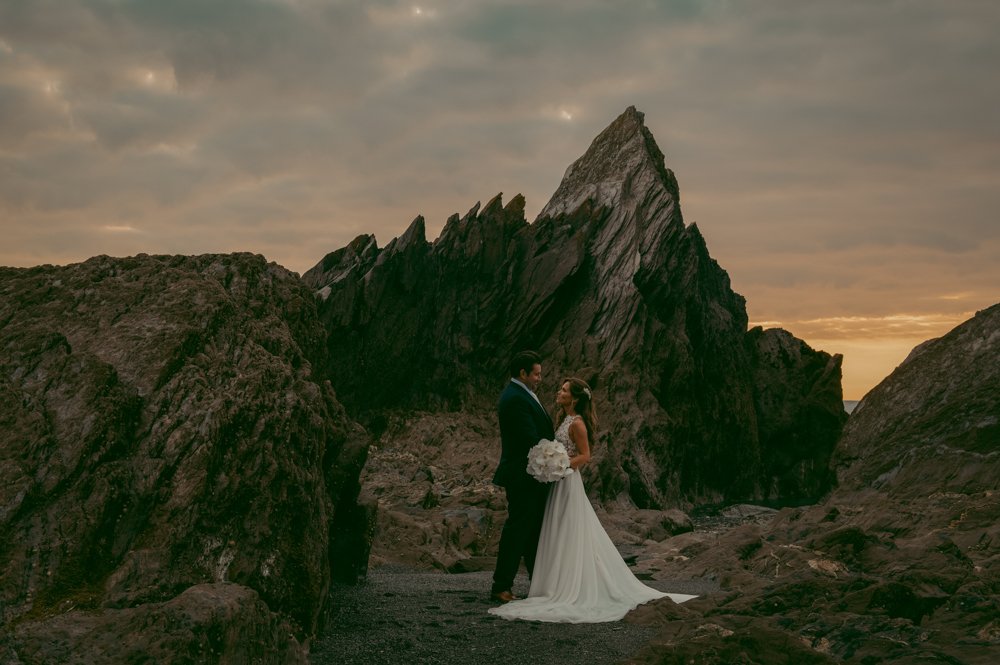 Bride and groom standing on rocky terrain with large jagged rocks and dramatic sky in the background.