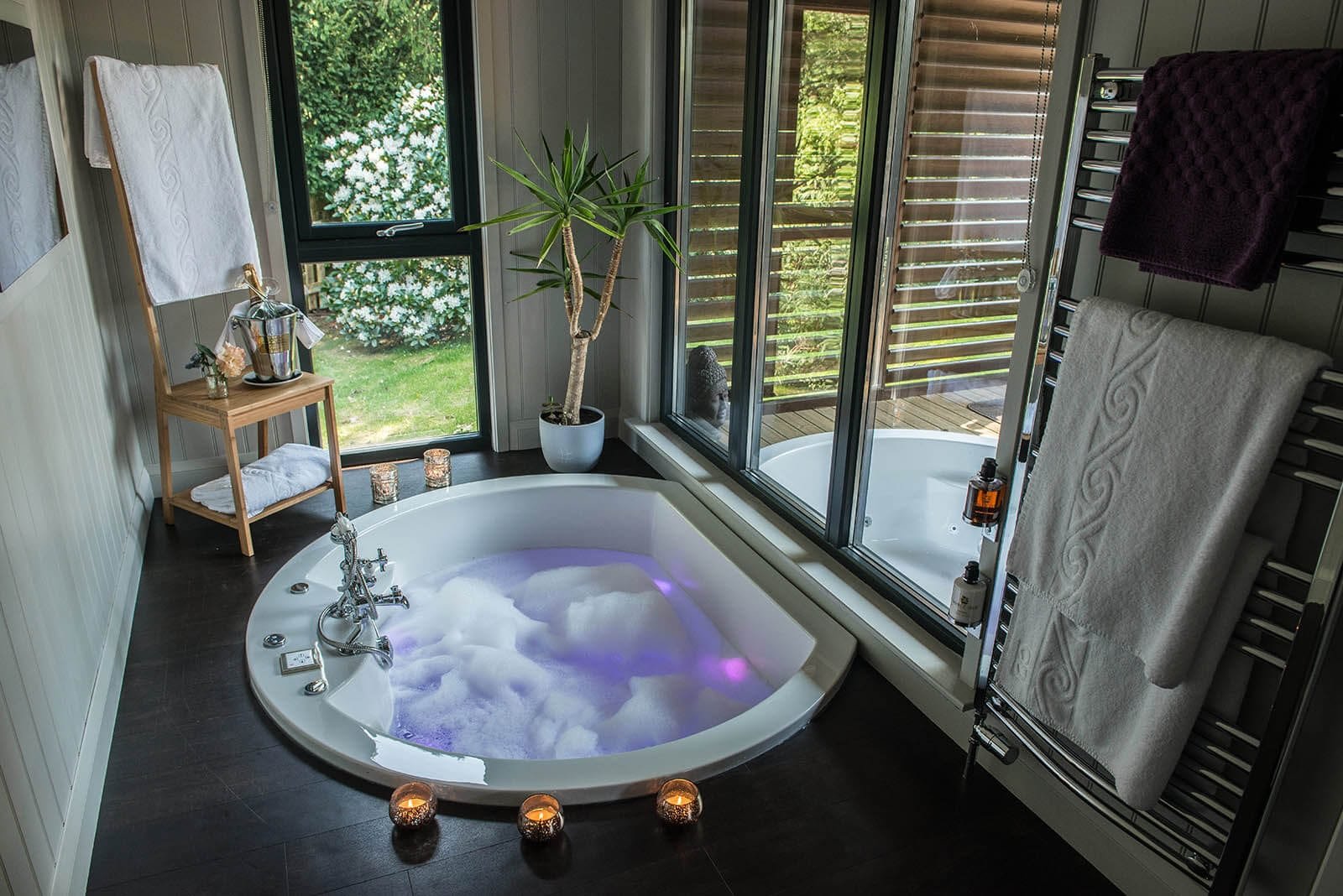 Modern bathroom with round bathtub, bubble bath, candles, plant, towel rack, and garden view.