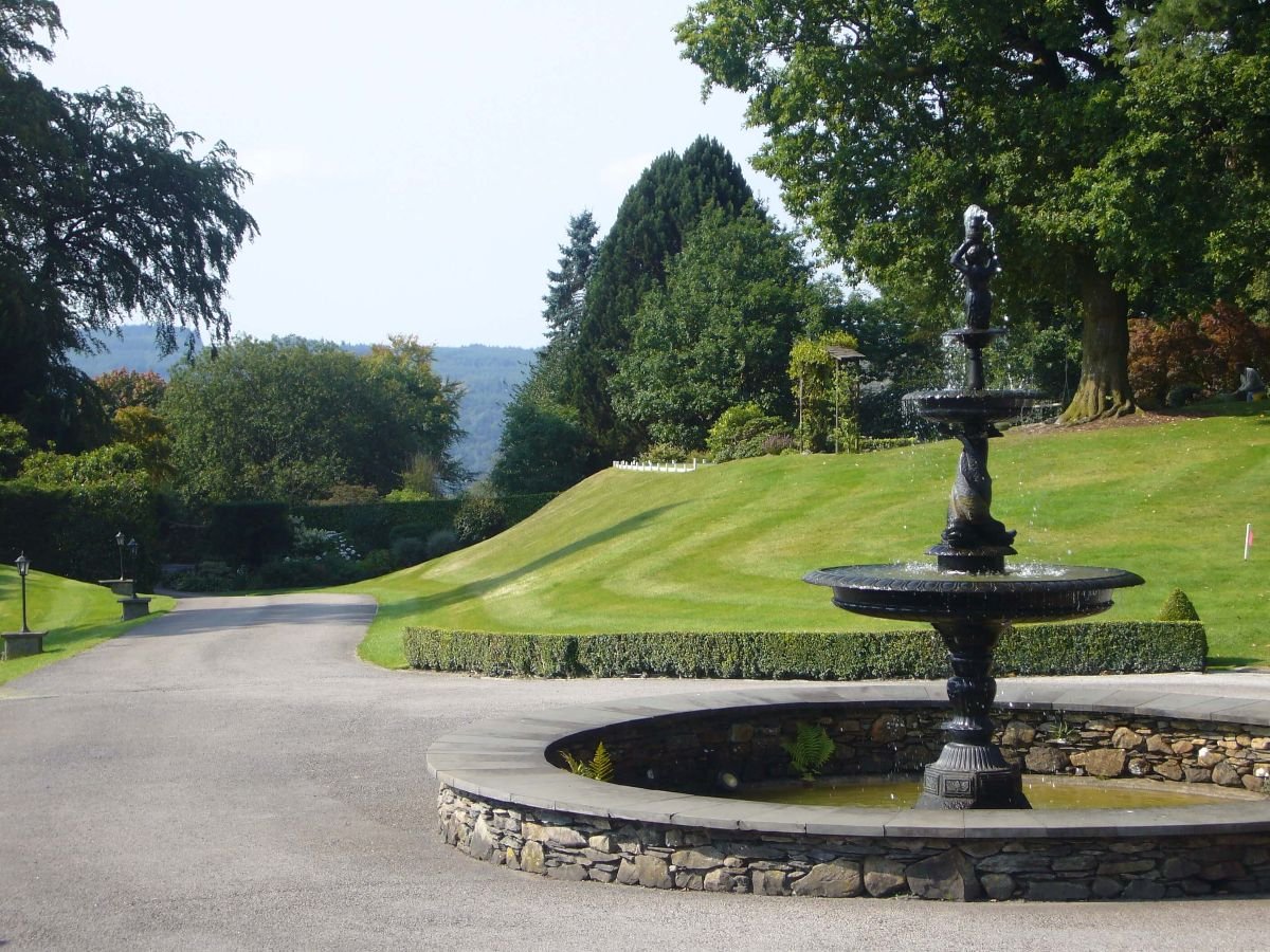 A landscaped garden with a paved path, a round stone fountain, manicured grass, and trees in the background.