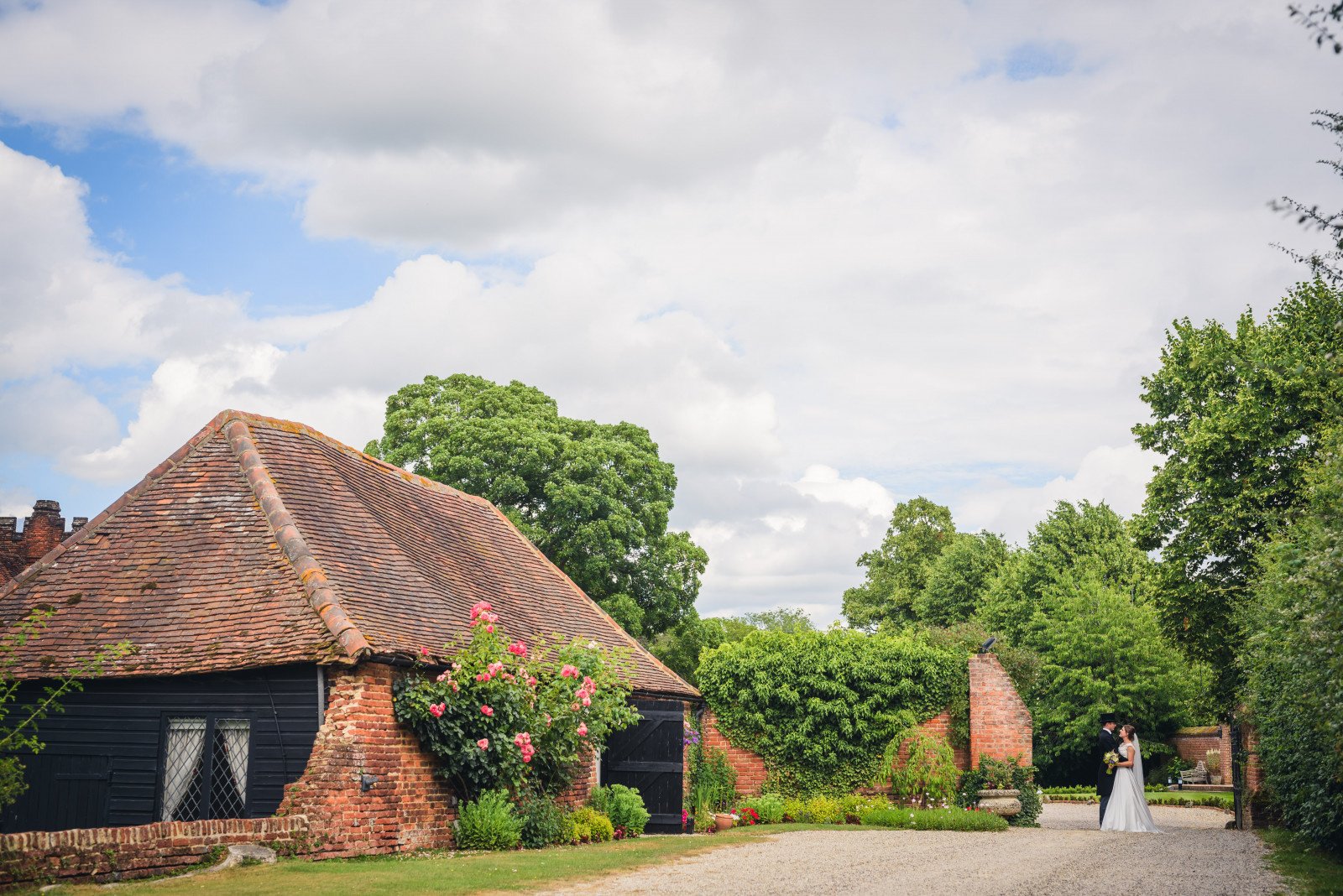 Rustic building with brick walls and a tile roof surrounded by greenery and flowers, with a couple standing in the distance on a gravel path under a cloudy sky.