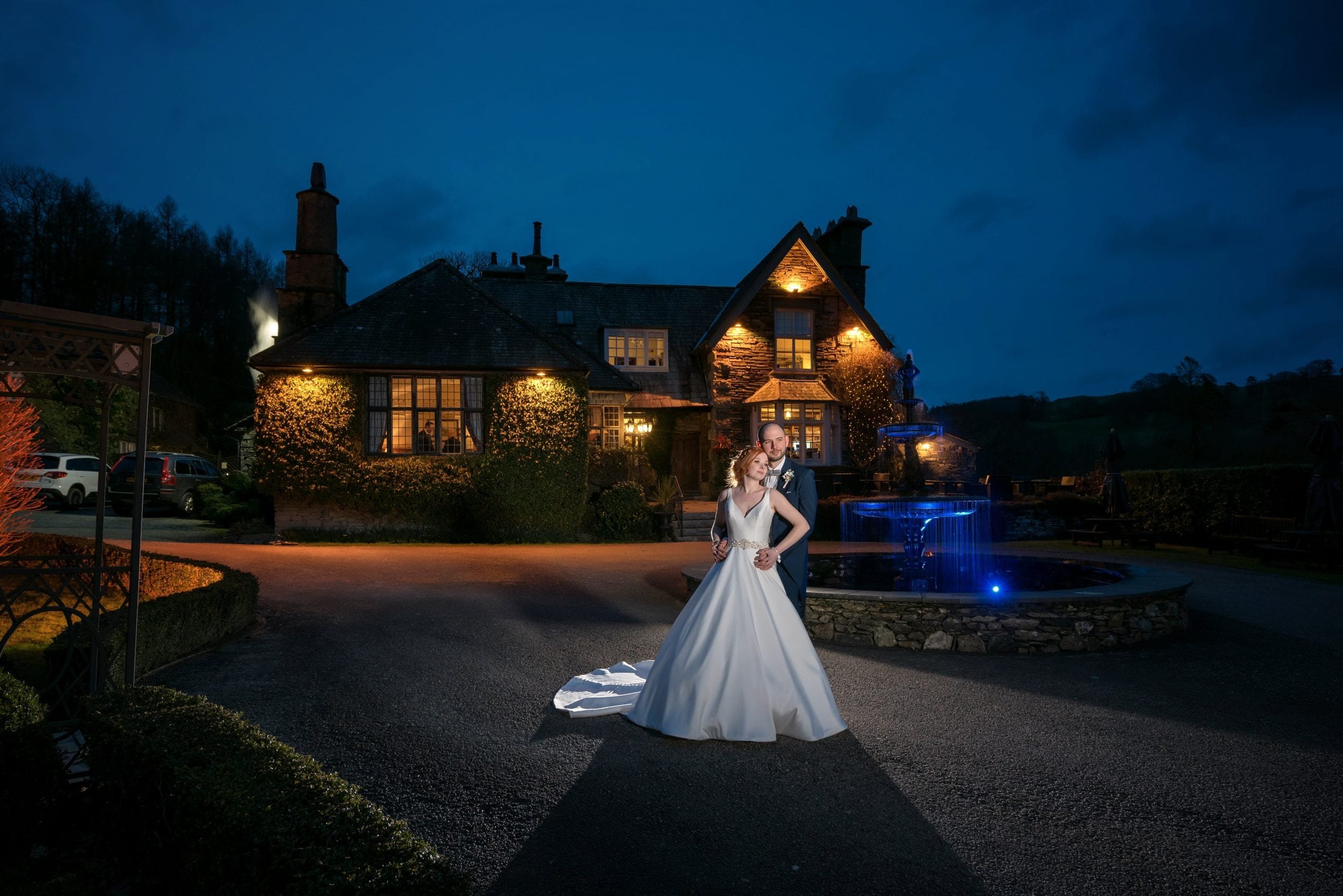 Bride and groom posing in front of a lit stone cottage with a fountain at night.