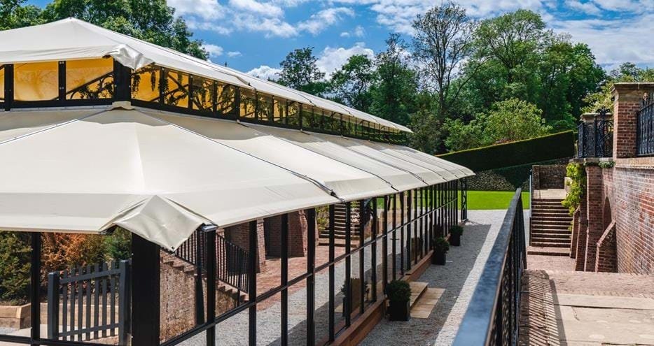 Outdoor walkway with large canopy and brick wall, surrounded by greenery and blue sky.