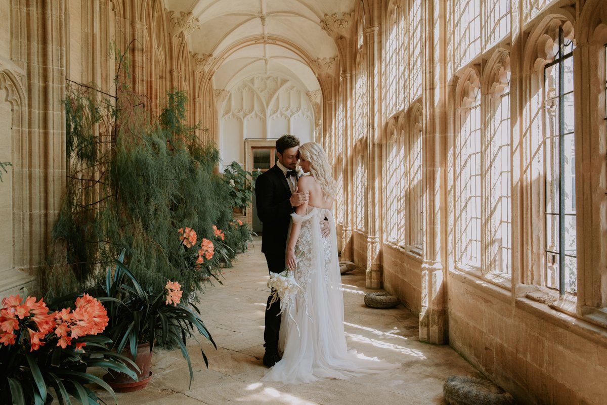 Bride and groom embracing in a sunlit corridor with Gothic architecture and flowers.