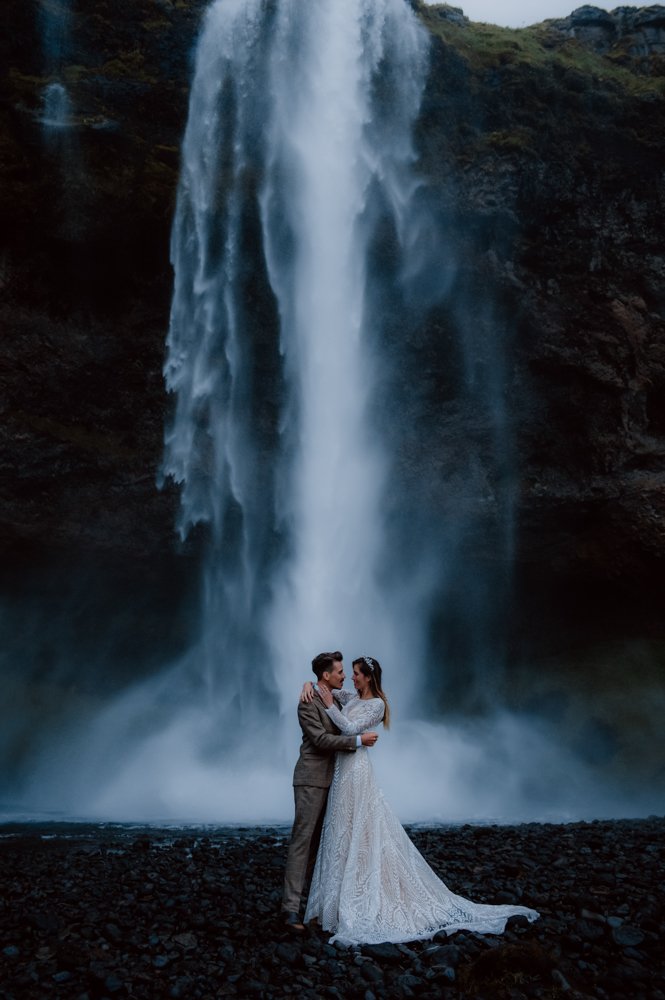 Bride and groom standing in front of a tall waterfall, dressed in wedding attire with the groom in a suit and bride in a lace gown.