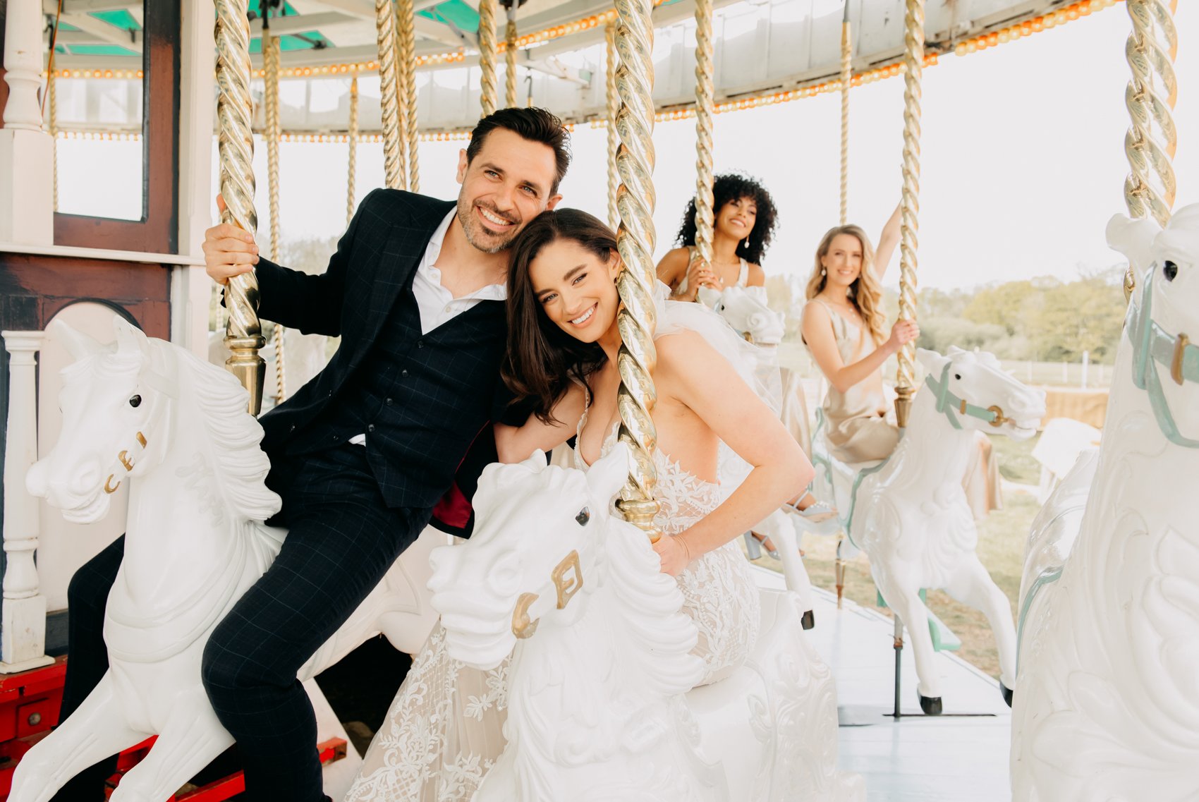Group of people smiling and riding carousel horses, with two in wedding attire and two in dresses.