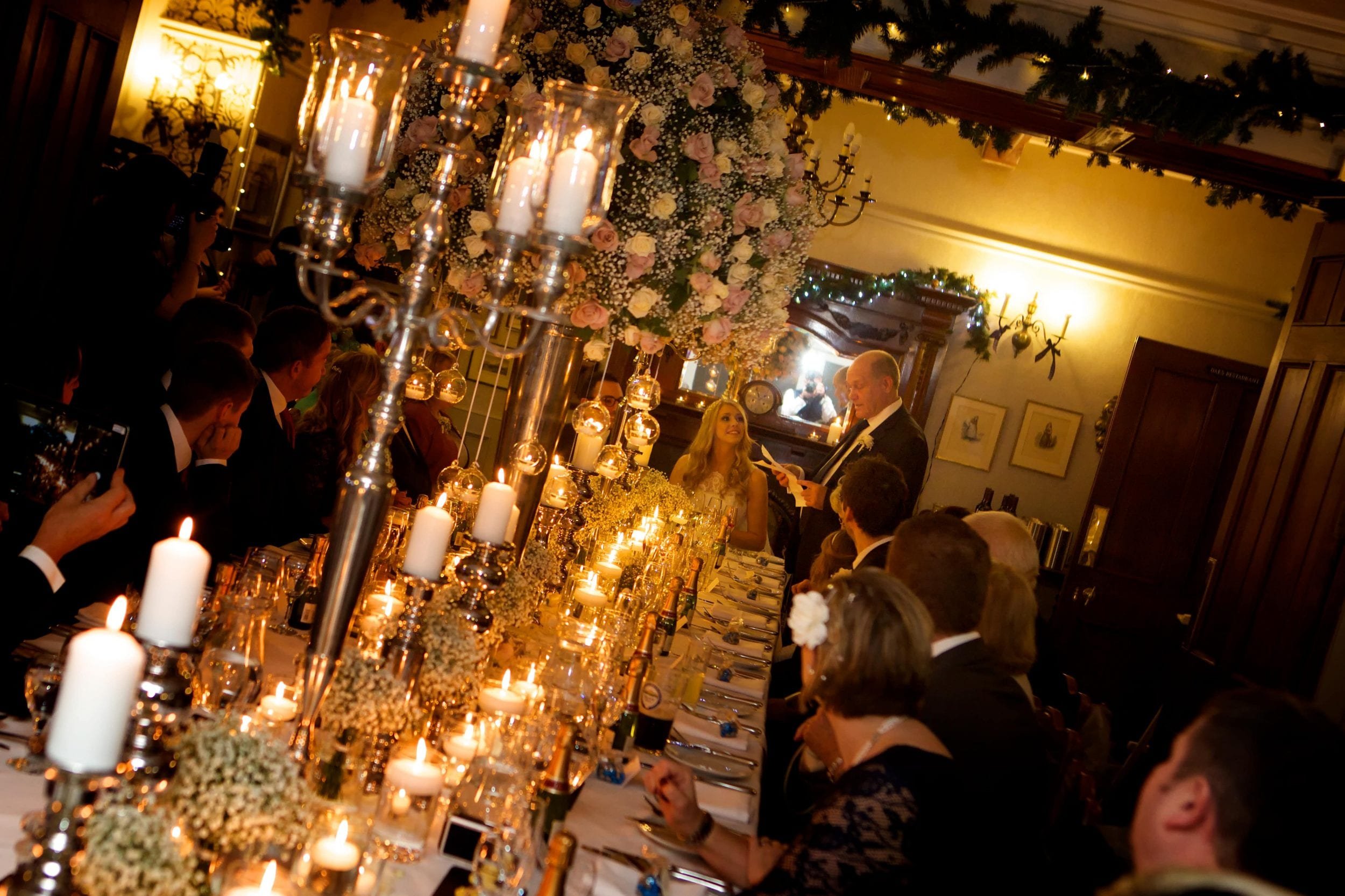 Elegant candlelit dinner party with guests seated at a long table adorned with floral arrangements and silver candelabras; a man stands, appearing to give a speech.
