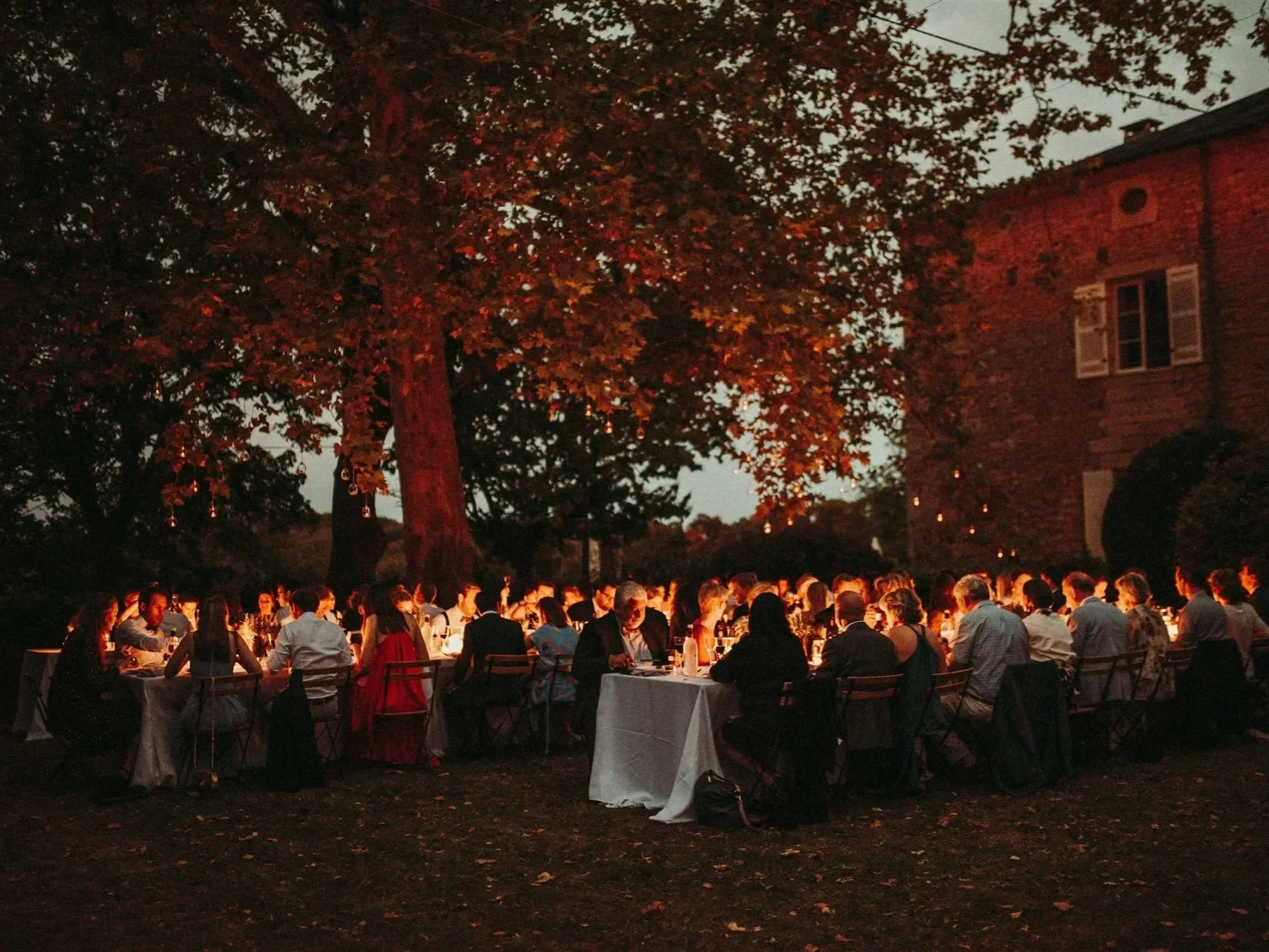 Outdoor evening dinner event with many people seated at long tables under large trees with autumn leaves, lit by candlelight, on a dark evening near a brick building.