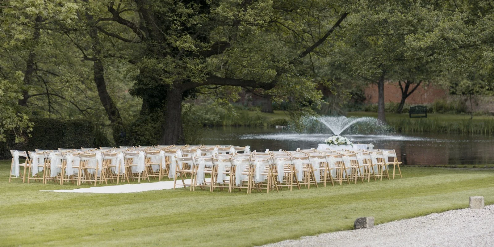 Outdoor wedding ceremony setup with wooden chairs draped in white fabric, facing a pond with a fountain and surrounded by trees.