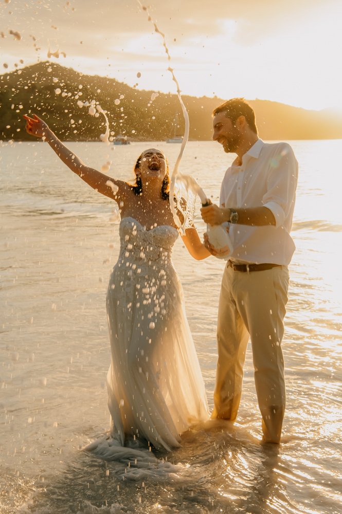 Bride and groom celebrating with champagne on a beach at sunset.
