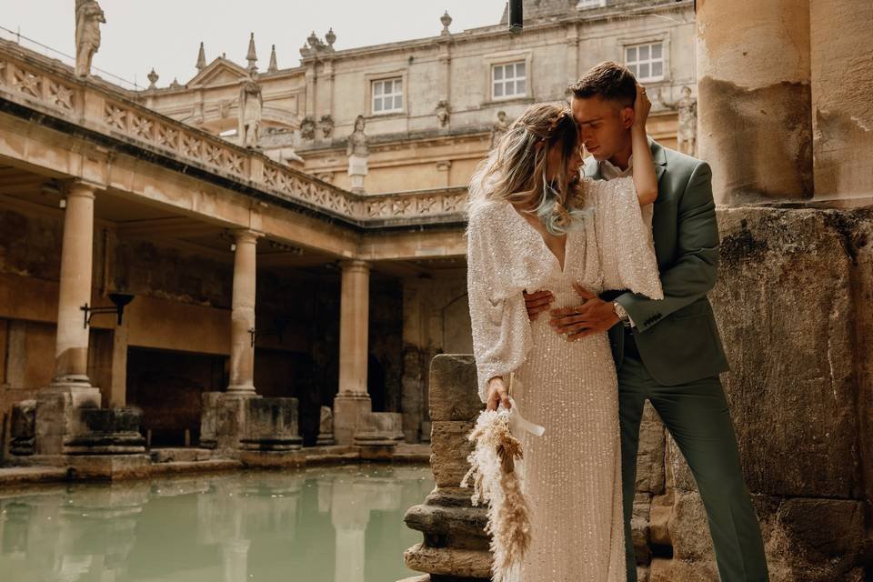 A couple in formal attire embraces romantically by an ancient Roman-style bath with columns and water.