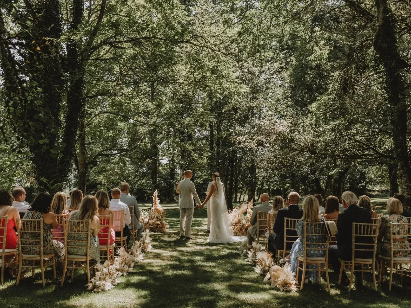 A wedding ceremony outdoors in a wooded area, with the bride and groom holding hands in front of seated guests.
