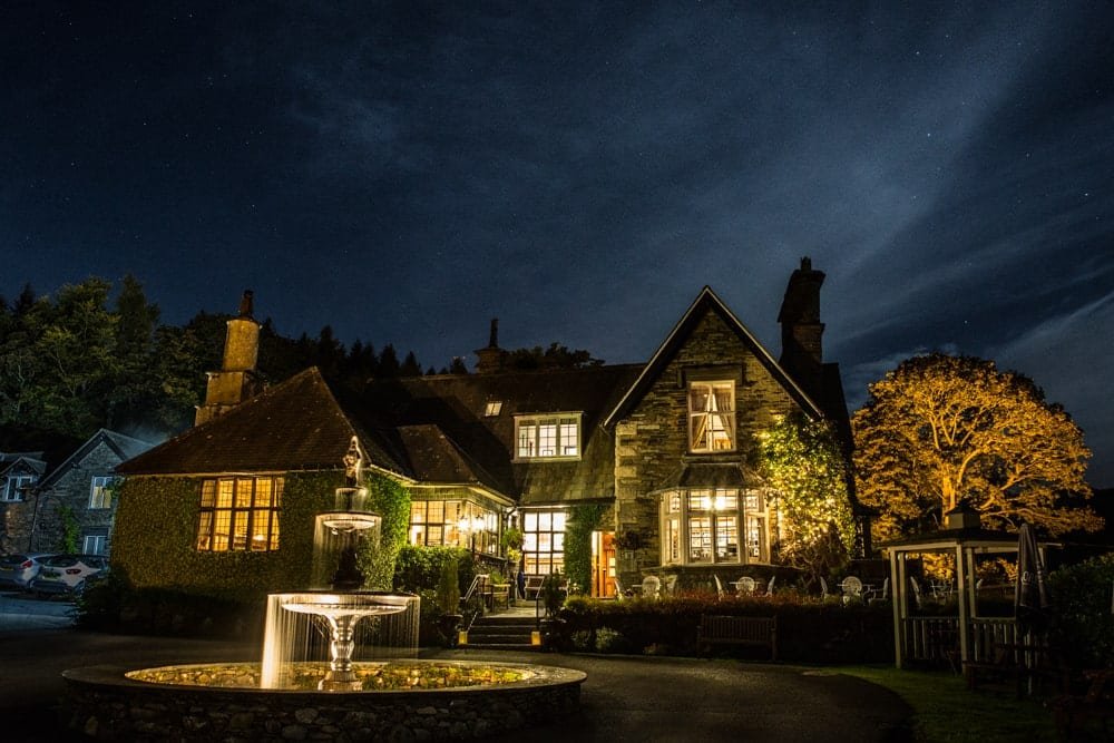 Night scene of a well-lit, ivy-covered stone house with a peaked roof and a fountain in the foreground, surrounded by trees.
