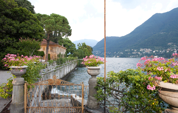Split view of a lakeside scene with pink flowers in containers, a stone building, and mountains in the background.