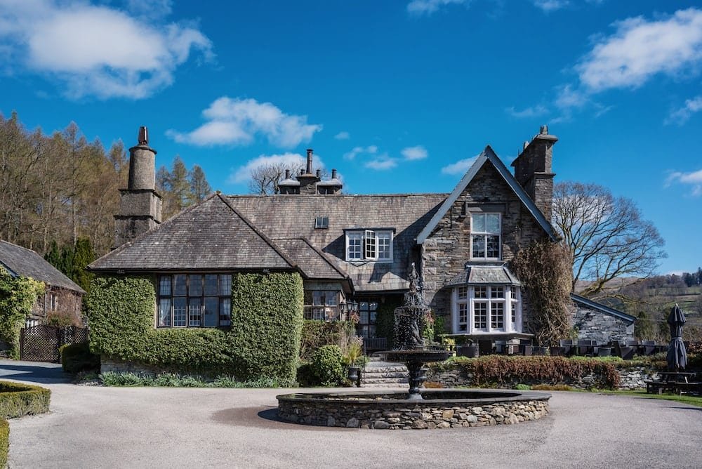 Country house with stone facade, ivy-covered walls, and a circular fountain in the foreground. Surrounded by trees under a blue sky with clouds.