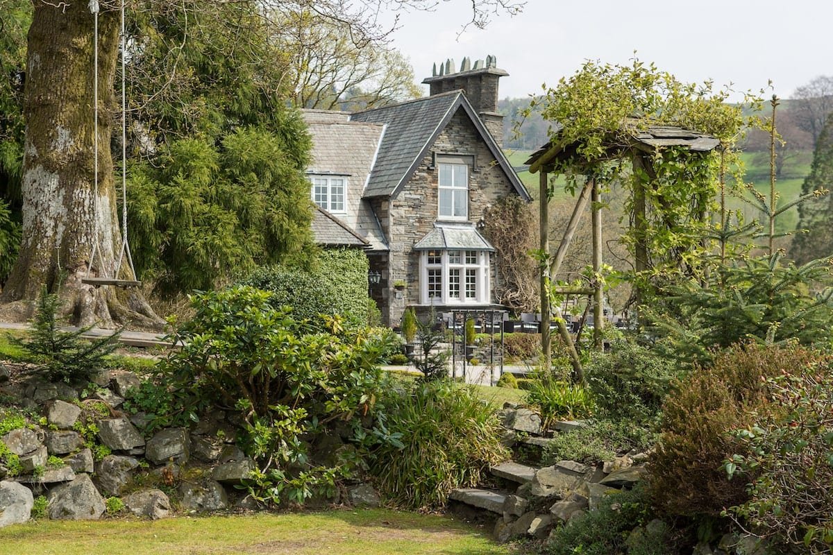A charming stone cottage surrounded by lush greenery and a garden. Features include a large tree with a swing, a patio area, and a trellis covered with plants. The setting appears to be tranquil and picturesque, ideal for a countryside retreat.