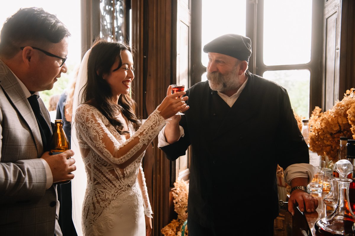Bride in lace gown toasting with chef, man in suit holding beer nearby, indoor celebration.