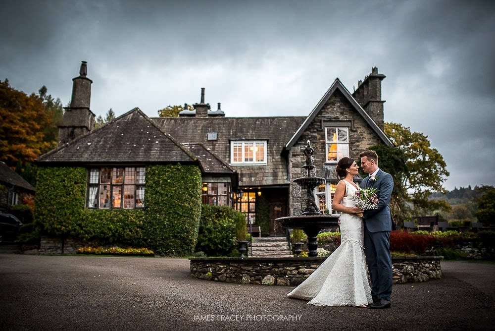 Bride and groom pose in front of an ivy-covered stone house with a fountain, surrounded by autumn trees.