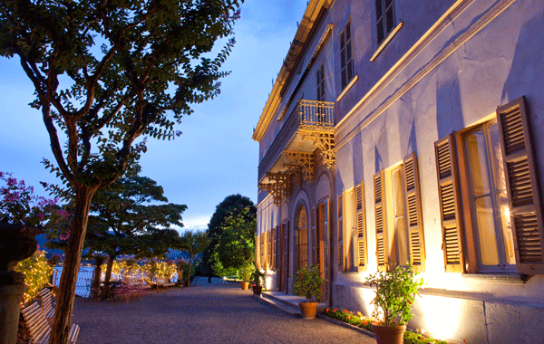 Elegant purple hotel with arched windows and wooden shutters, illuminated by warm lighting at dusk, with lit trees and a gravel pathway.