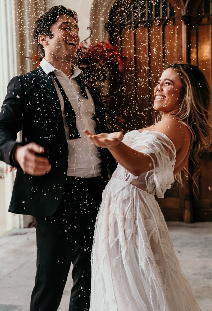 Bride and groom celebrating with confetti, smiling and joyful in wedding attire.