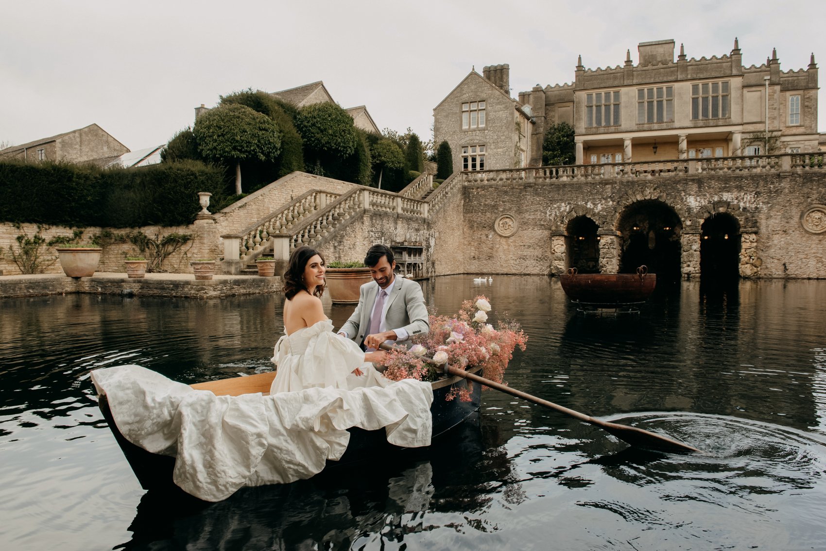 A couple in formal attire rowing a boat on a serene pond near a historic building, decorated with flowers.