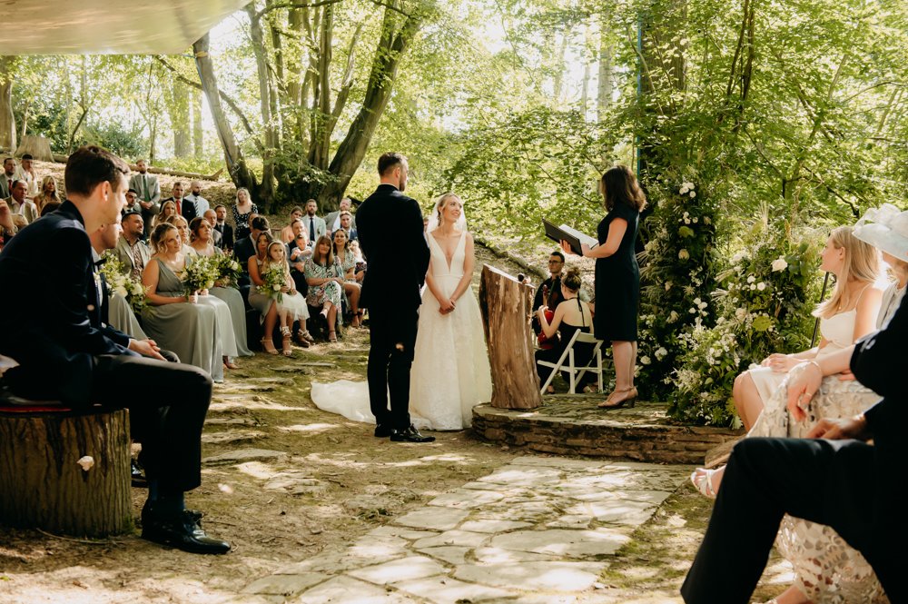 Outdoor wedding ceremony with a bride and groom standing before an officiant in a forest setting, surrounded by guests seated on log benches.