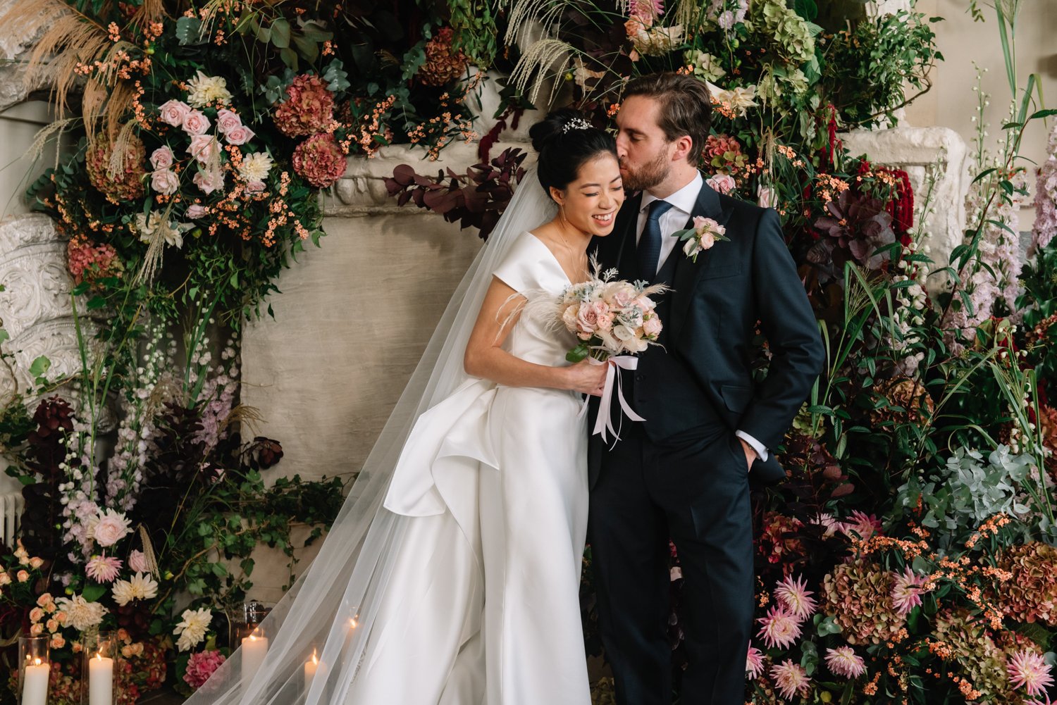 Bride and groom in wedding attire embracing, surrounded by colorful floral arrangements and lit candles.