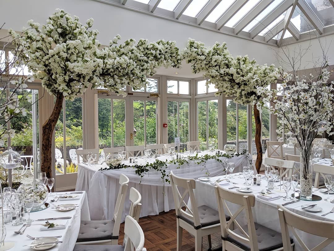 Elegant wedding reception setup with white flower arrangements, tables set with white linens and glassware, surrounded by greenery in a bright conservatory with glass ceilings.