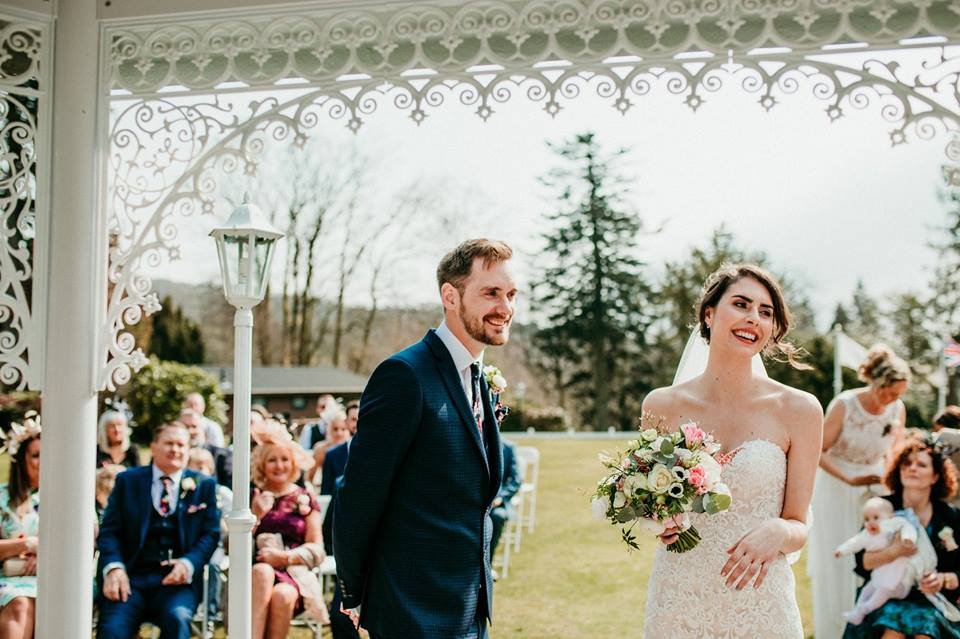 A bride and groom standing under a decorative archway during an outdoor wedding ceremony. The bride is holding a bouquet, and guests are seated behind them.