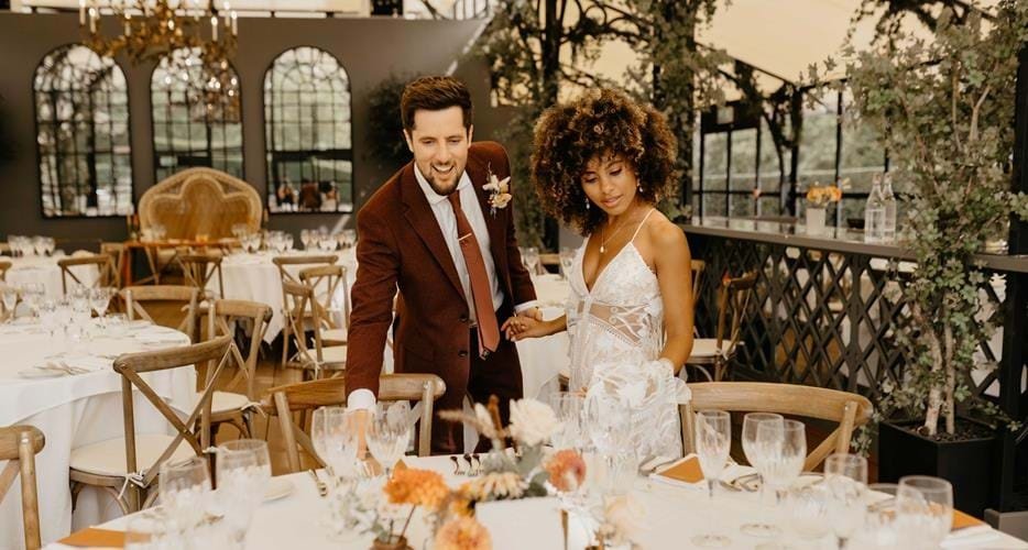 A couple dressed in wedding attire is arranging tables at an elegantly decorated venue. The setting includes round tables with white tablecloths, floral centerpieces, and empty wine glasses. The background has large windows and greenery, creating a s