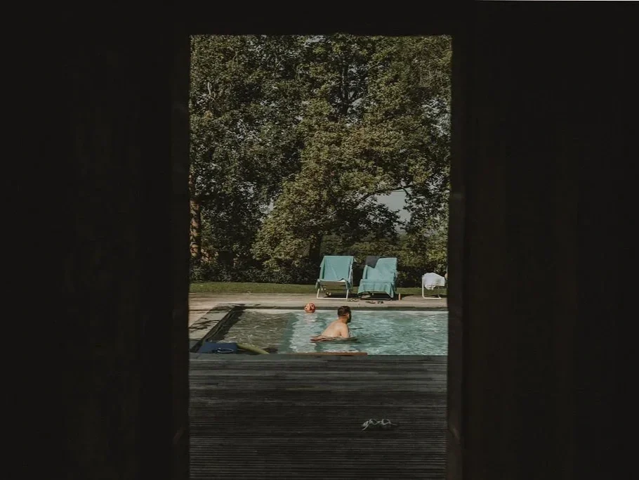 A person swimming in an outdoor pool, viewed through a dark window or opening with trees and lounge chairs in the background.