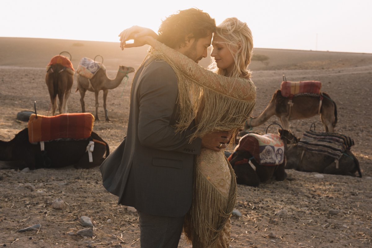 A couple embracing in a desert landscape with camels carrying colorful saddles in the background.