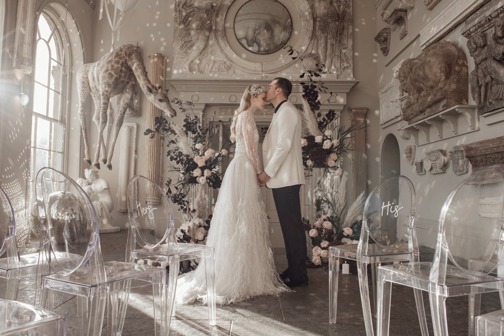 Couple in wedding attire kissing in a decorated room with glass chairs, floral arrangements, and a giraffe statue.