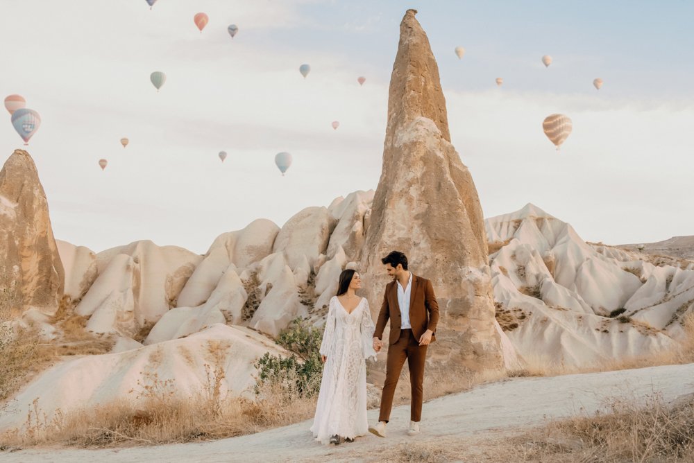 A couple in formal attire walking in Cappadocia, with unique rock formations and hot air balloons in the background.