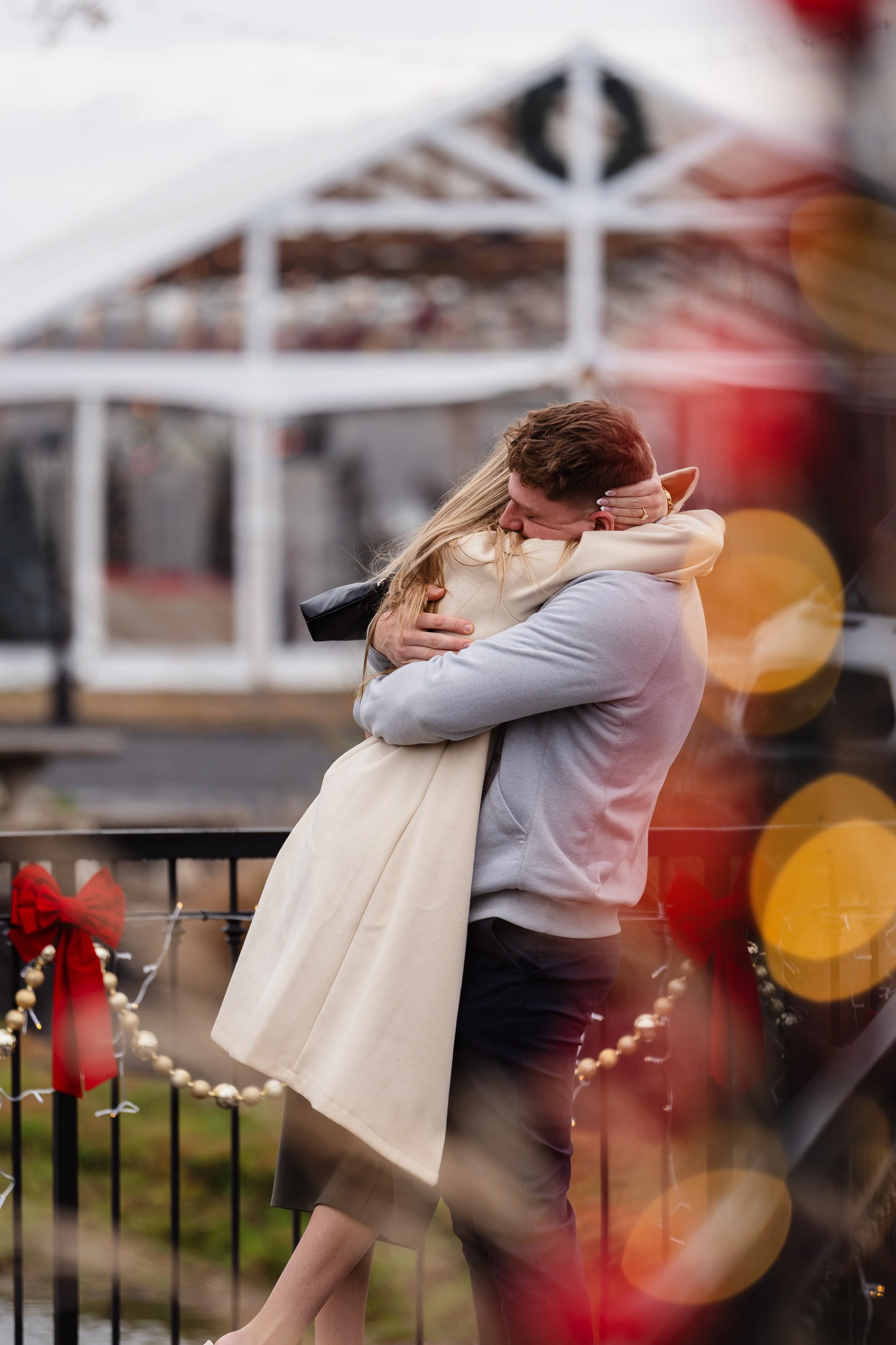 A couple embracing each other outdoors during proposal, with festive decorations and a blurred background at Renault Winery in Egg Harbor, New Jersey.