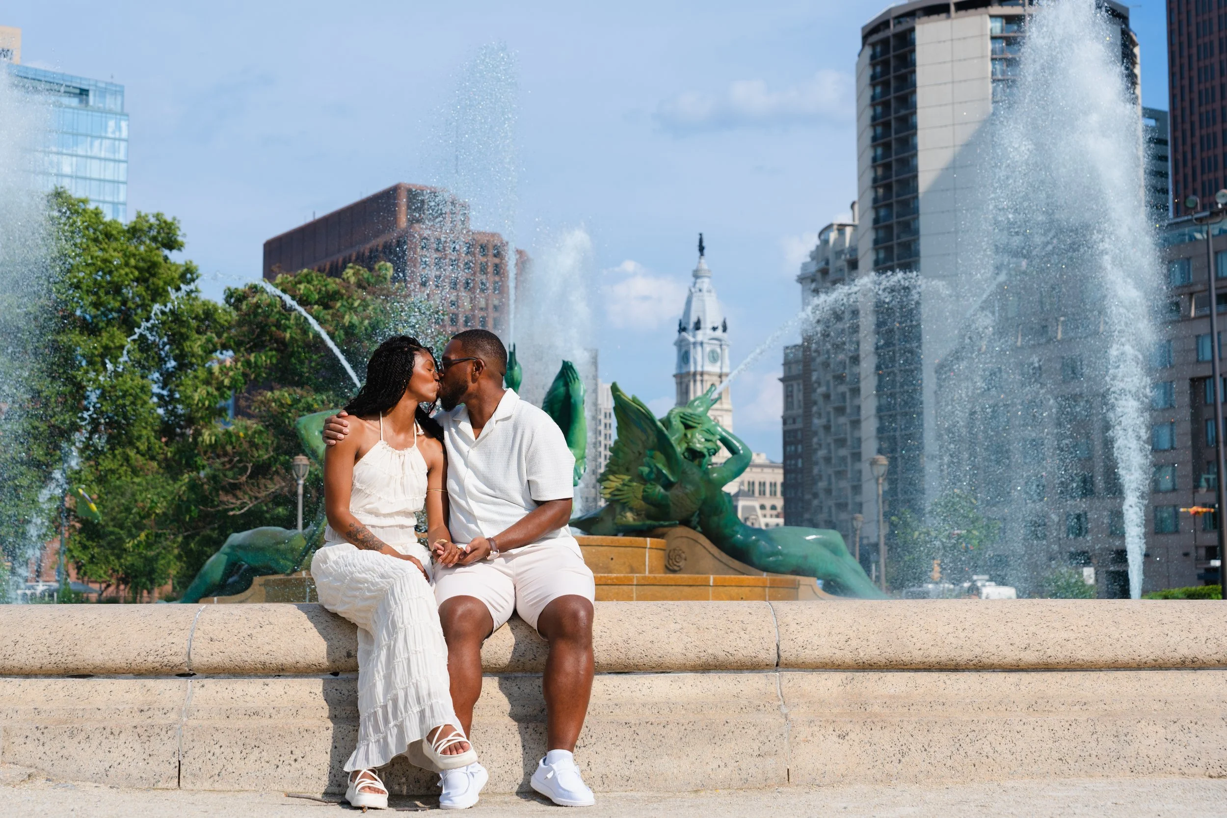 A couple sharing a kiss while sitting on a fountain in a city park with tall buildings and a clock tower in the background. Logan Square, Philadelphia, PA.