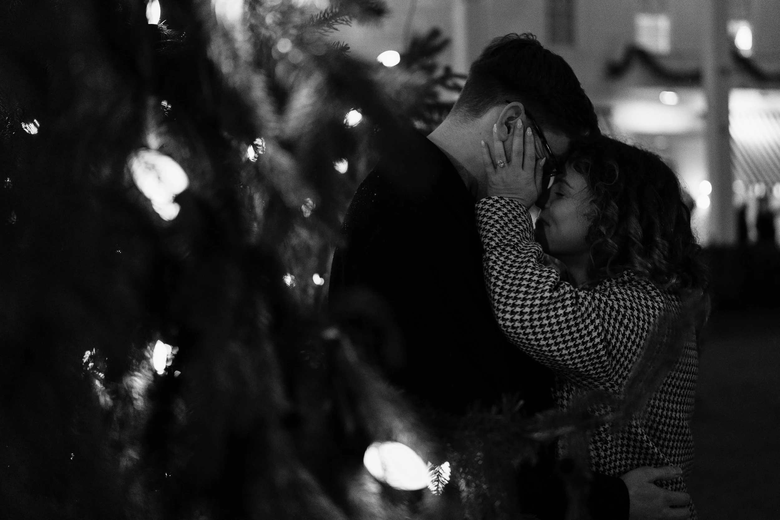 A couple sharing an intimate moment, holding each other close, with the woman’s face cupping the man’s face, surrounded by blurred lights and a Christmas tree in the foreground, for Propsal photo shoot at Congress Hall in Cape May, New Jersey.