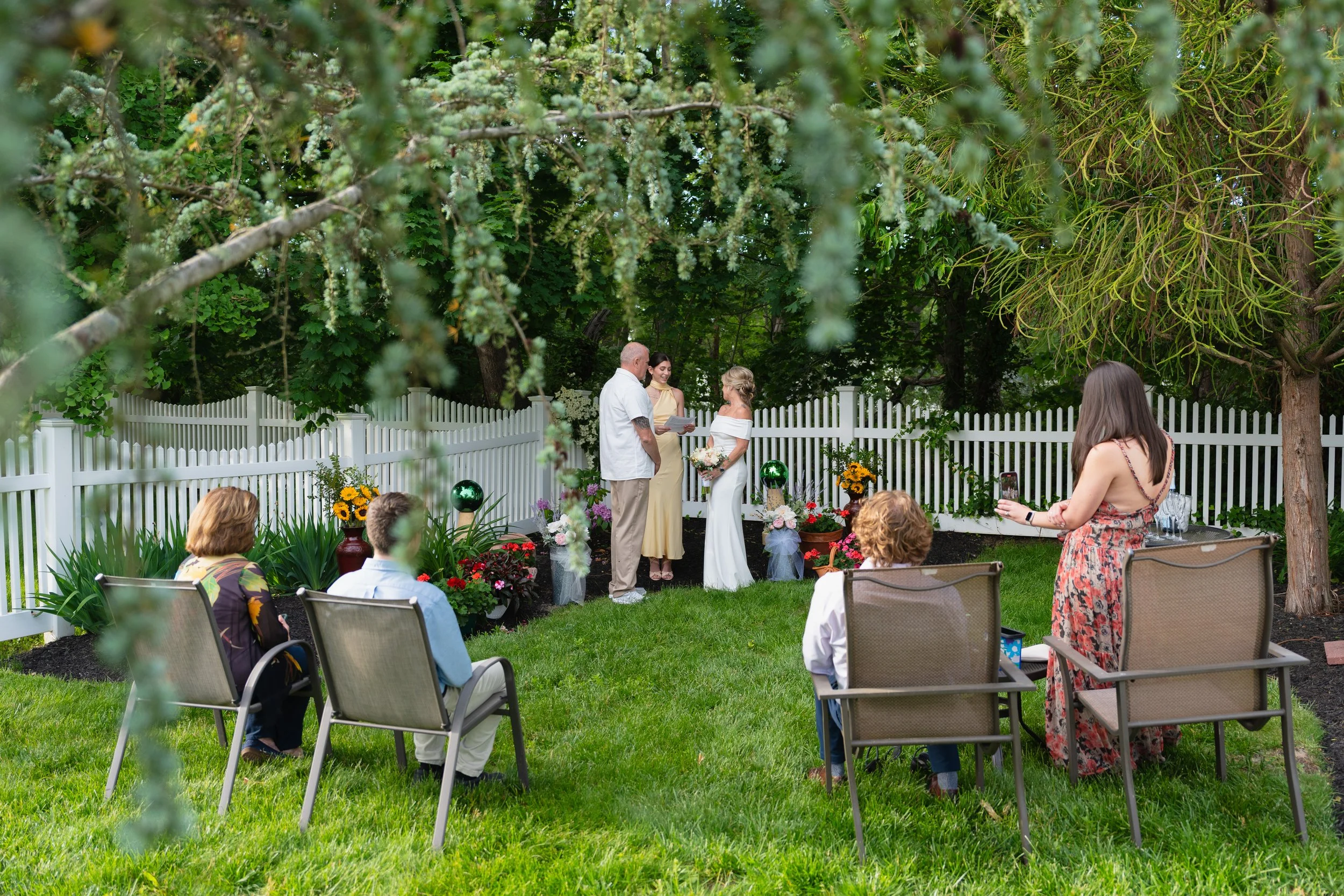 Outdoor wedding ceremony in a garden with a white picket fence. A bride and groom stand in front of an officia, exchanging vows. Three guests sit on chairs while a woman is taking a photo. The scene is decorated with flowers, plants, and green trees.