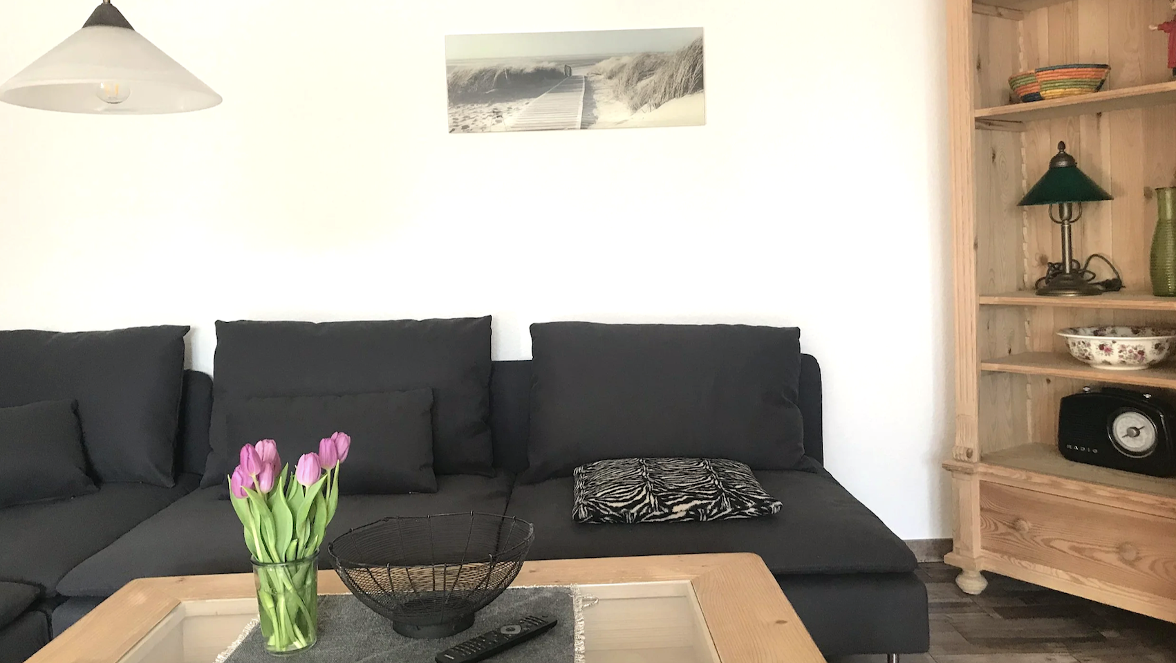 Wohnzimmer mit schwarzer Couch, einem Zebra-Muster-Kissen, einer Holzwandregal mit Lampen, Vasen, Schüssel und Radiowecker. Es gibt eine Vase mit pinken Tulpen auf dem Couchtisch und eine Wanddekoration mit Strandweg. Ein Deckel an der Decke.