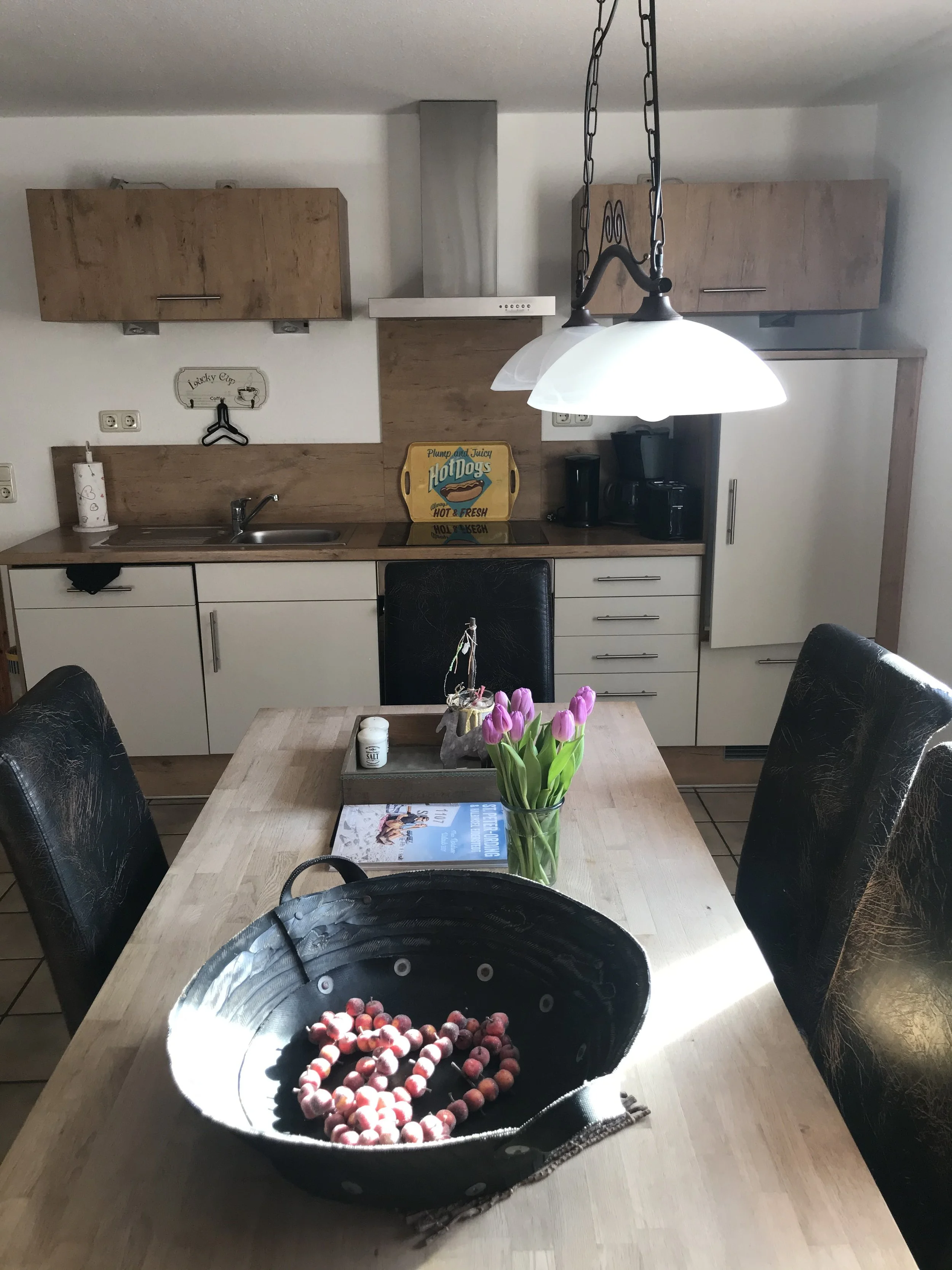 Ein Esszimmertisch mit schwarzen Stühlen, einem Korb mit roten Beeren, einer Vase mit pinkfarbenen Tulpen, und einer Zeitschrift, im Hintergrund eine Küchenzeile mit Holz- und weißen Schränken, auf der die Tafel steht.