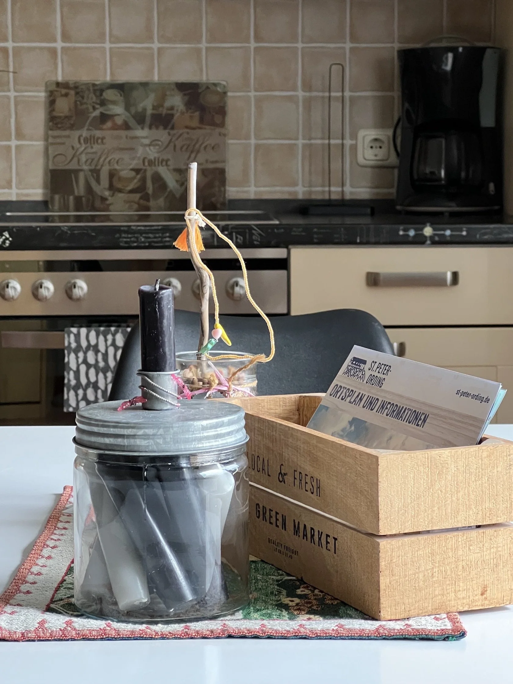 Küchenszene mit Kerze in einem Glas, einer Holzbox mit Informationsmaterial, im Hintergrund Herd und Kaffeemaschine.