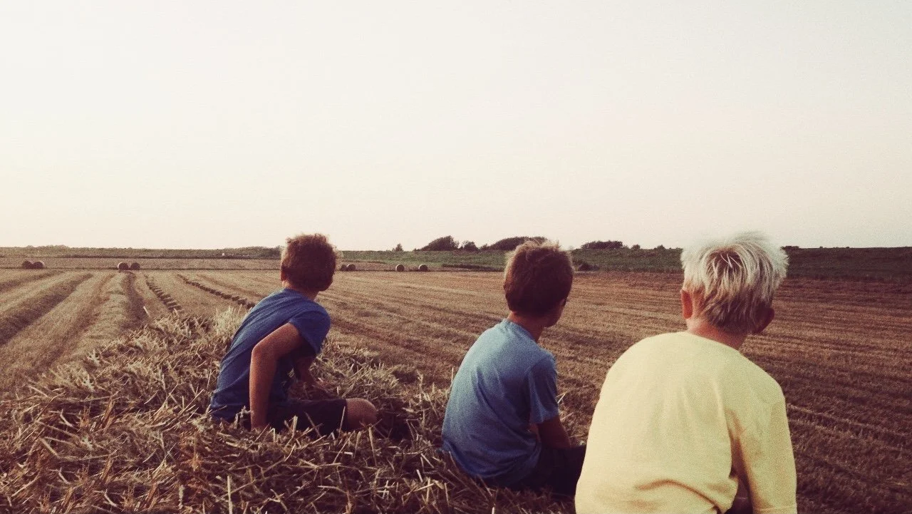 Drei Jugendliche sitzen auf einem Heuhaufen und schauen auf ein Feld mit Strohballen im Sonnenuntergang.